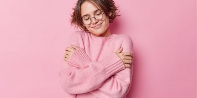 photo-tender-pretty-woman-with-short-hairs-embraces-herself-feels-comfortable-new-soft-jumper-wears-round-spectacles-vision-correction-isolated-pink-background-self-care-concept